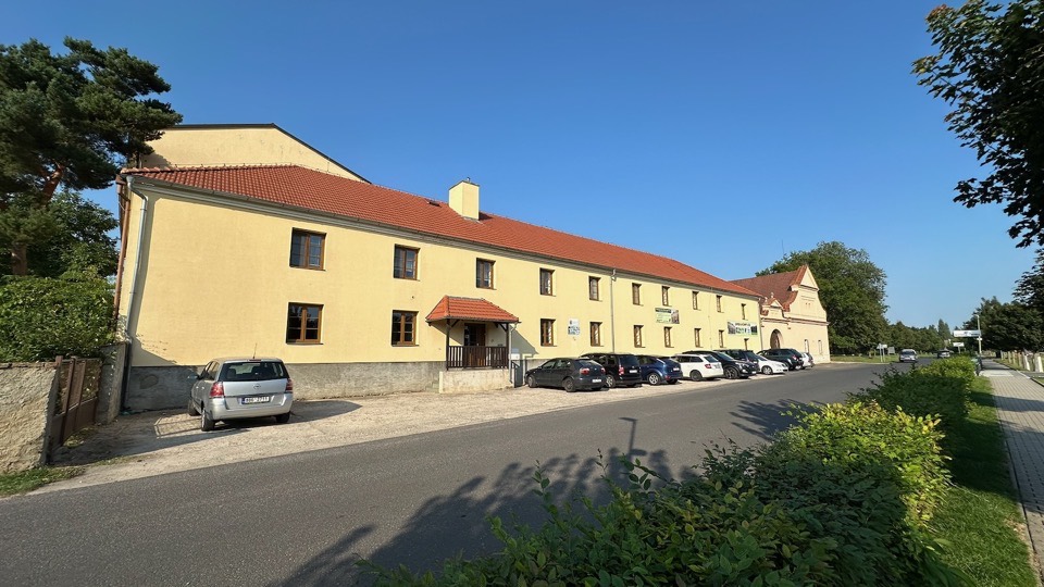 Stará budova ubytovny podél silnice, v pozadí historická brána areálu